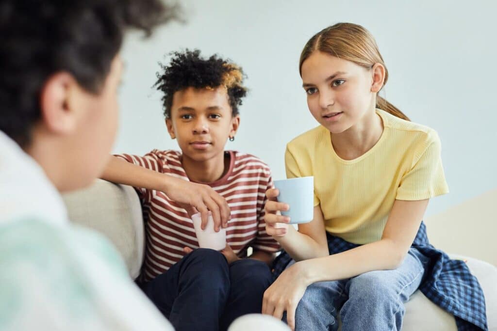 Group of positive diverse children sitting on sofa and holding cups of drink while spending time together against white wall