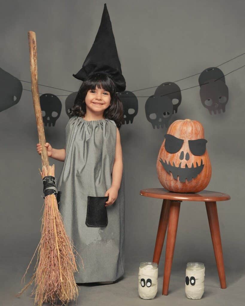 girl in grey dressed as a witch standing beside jack o lantern